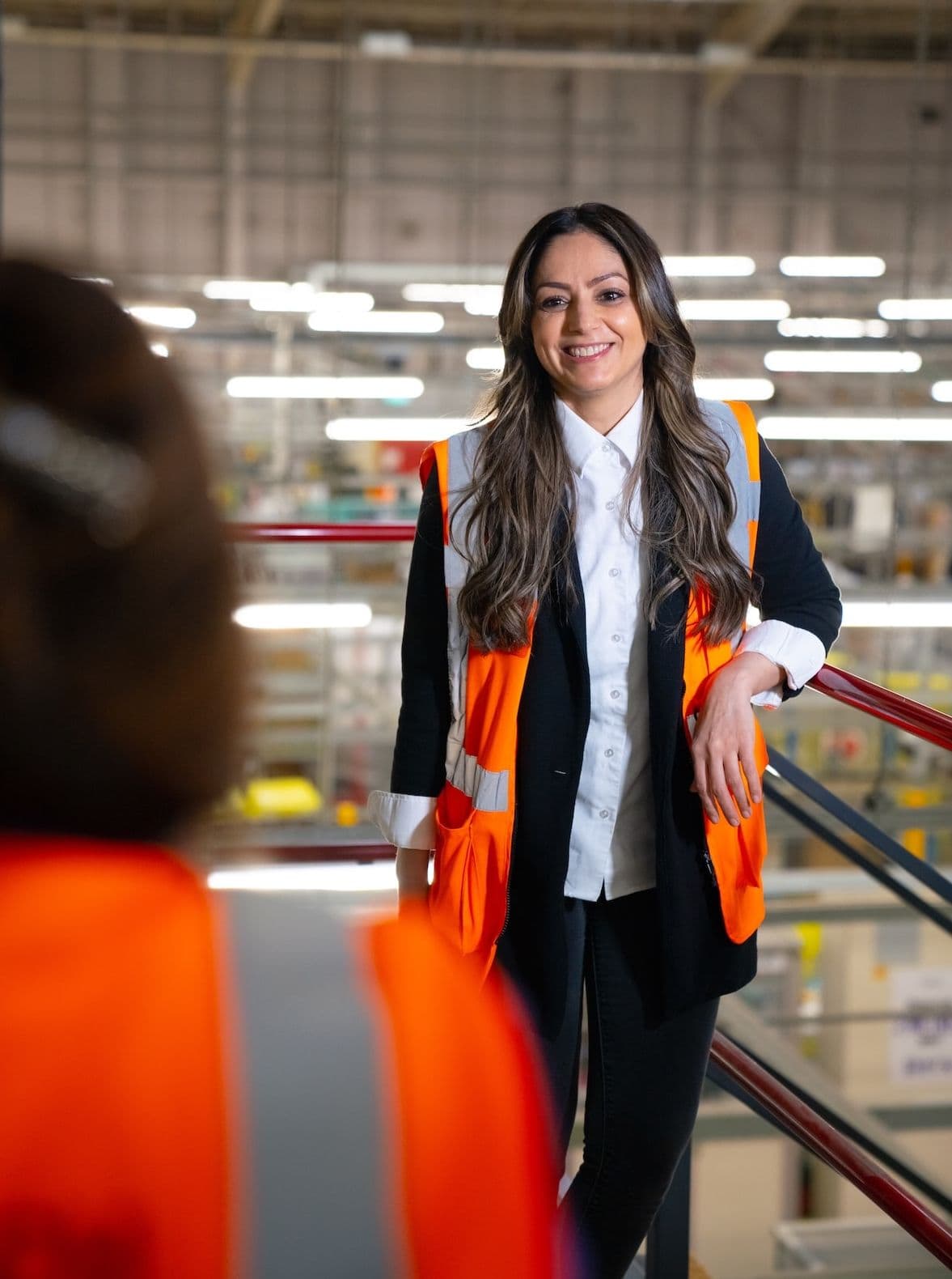 Woman in an orange safety vest smiling during a conversation in a brightly lit warehouse setting.