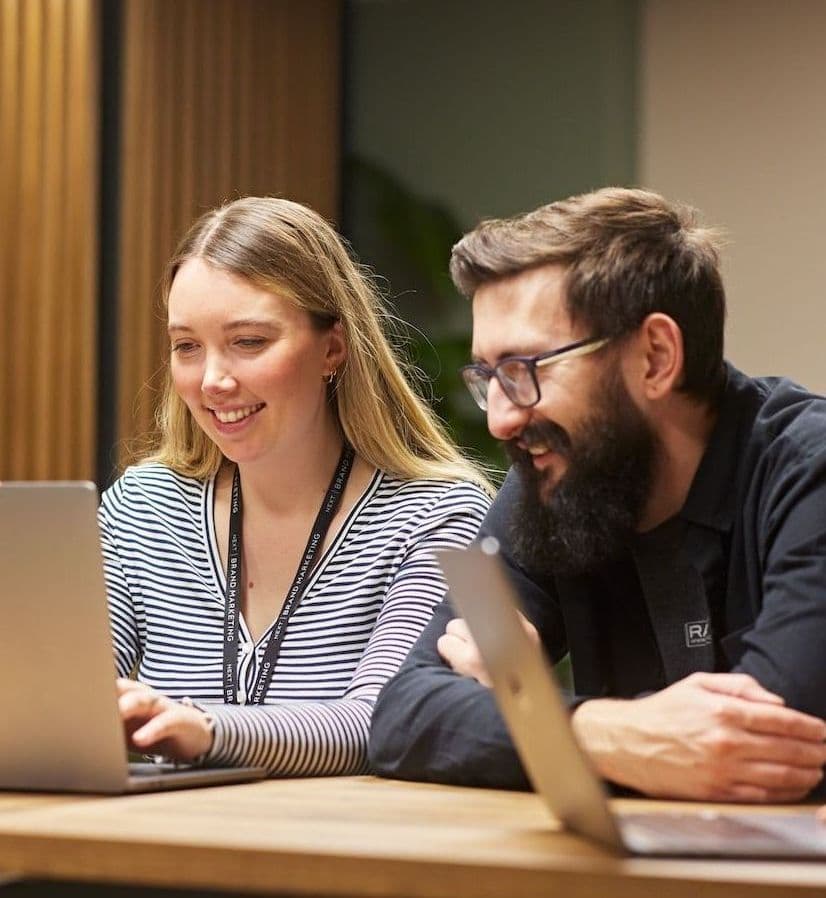 Two colleagues smiling and working together at laptops in a modern meeting space.