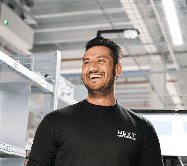 Smiling employee wearing a “NEXT Distribution” shirt, standing in a warehouse with numbered metal shelves.