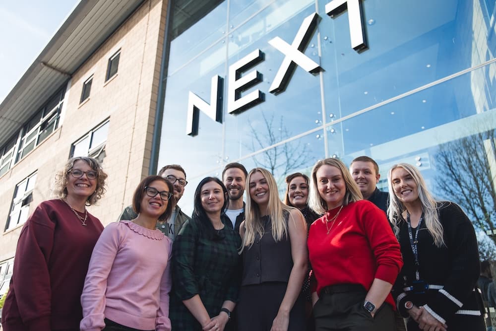 A group of smiling NEXT employees stand together outside the company’s modern glass-fronted building, posing beneath the large NEXT logo.