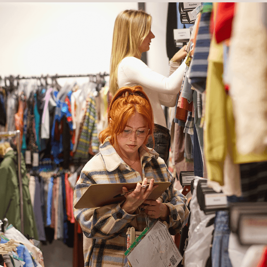Two women working in a clothing storage, one writes on a clipboard while the other selects garments from a rack.