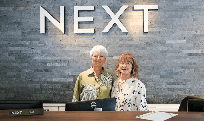Two women standing behind a reception desk with "NEXT" displayed on the wall behind them, both smiling toward the camera.