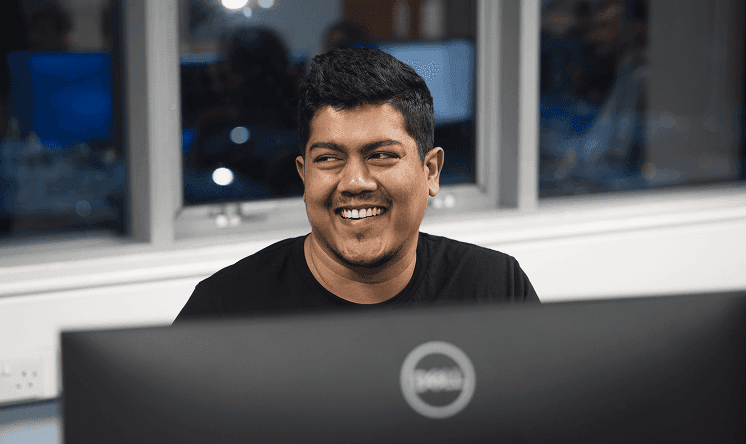 Man smiling while working at a computer in an office setting, with a window and colleagues visible in the background.