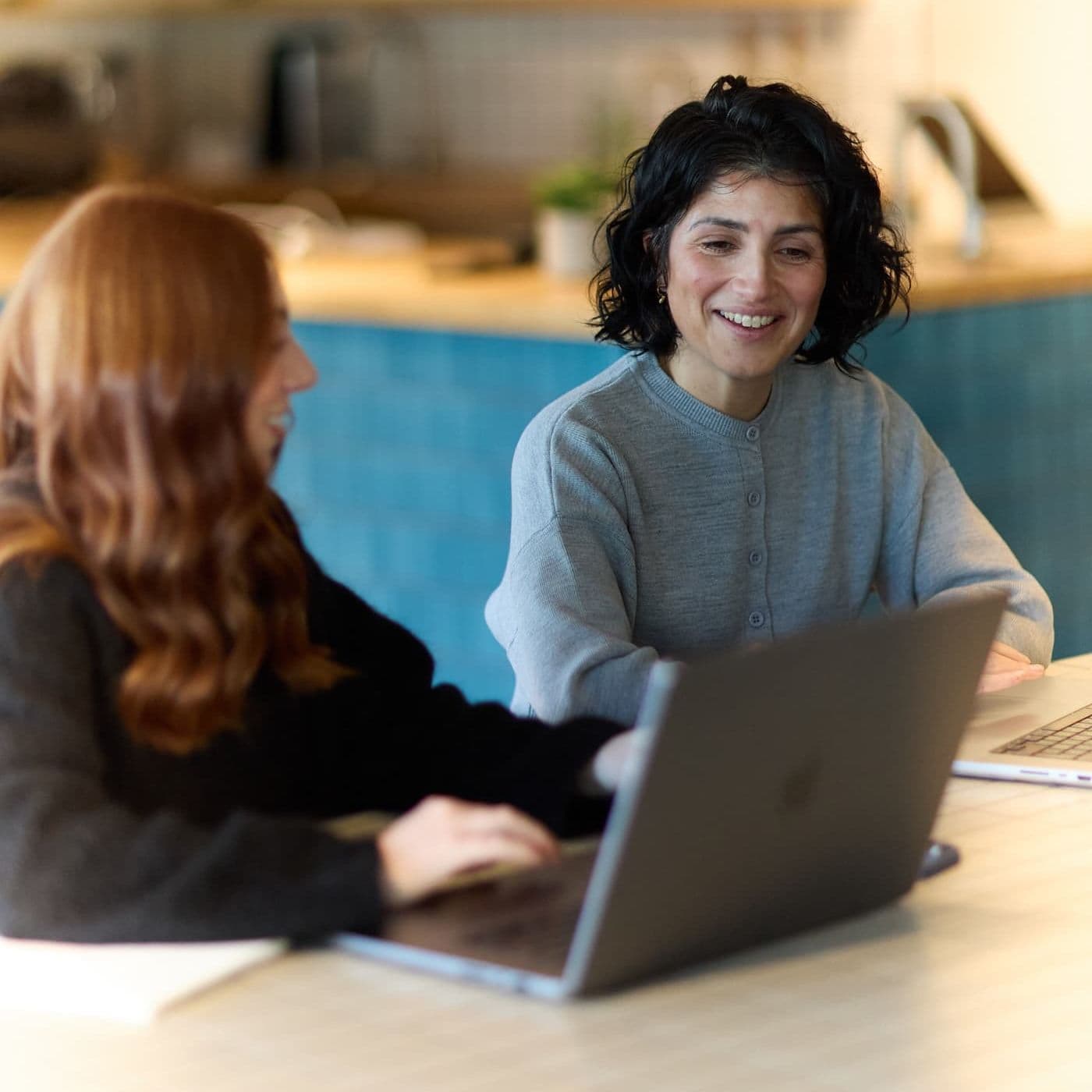 Two colleagues smiling and working together at laptops in a modern, plant-filled workspace.