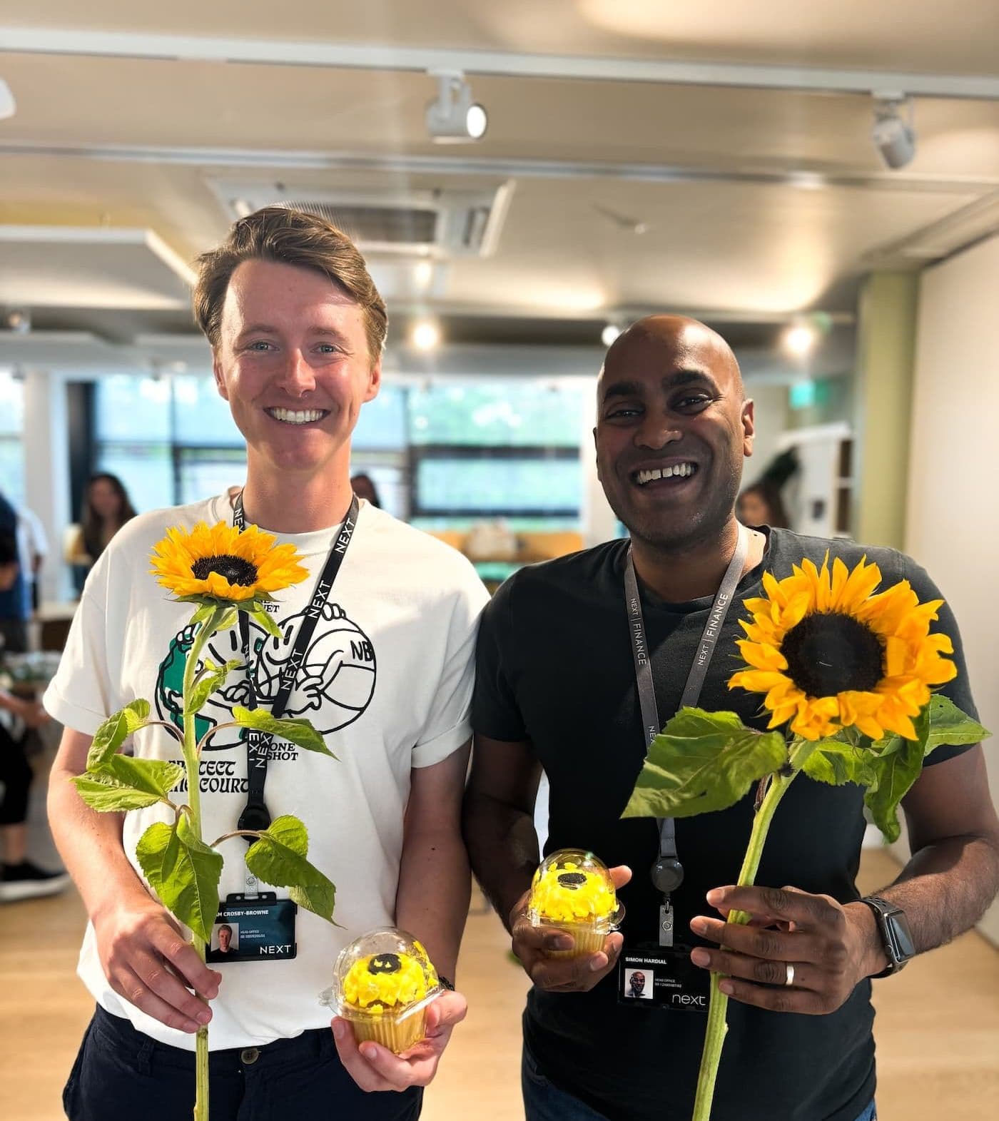 Two colleagues are smiling and holding sunflowers and sunflower-themed cupcakes, wearing NEXT lanyards indoors.