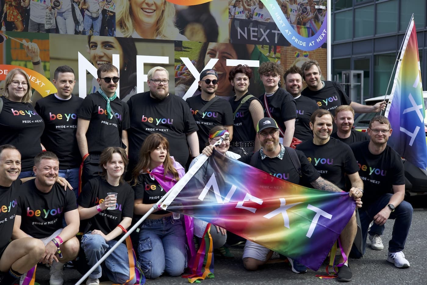Group of NEXT employees in 'be you' Pride shirts holding branded rainbow flags at a Pride event.