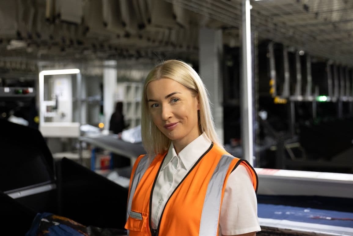 Woman in an orange high-visibility vest smiling in a warehouse or distribution centre setting.