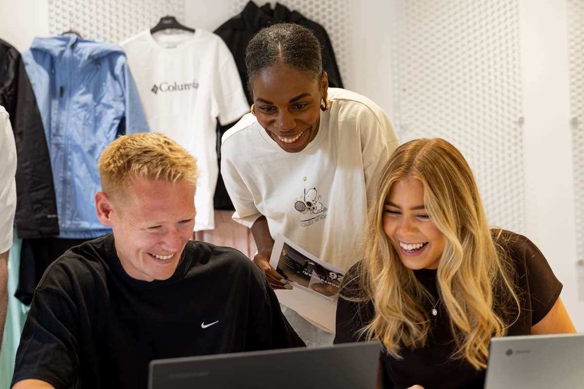 Three colleagues laughing and working together on laptops in a retail space with branded clothing in the background.