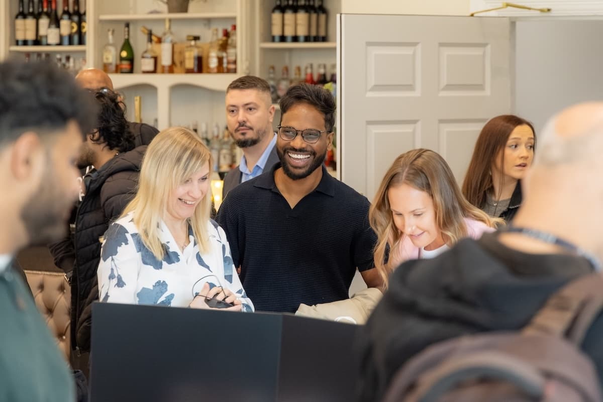 Group of people chatting and smiling around a table at a social or networking event, with bottles displayed in the background.