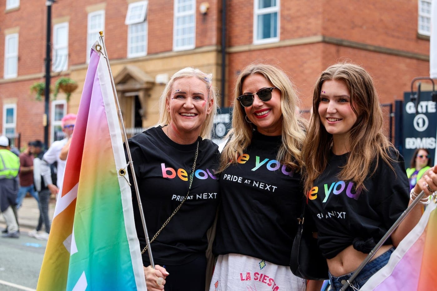 Three people smiling and posing at a Pride parade wearing “be you – PRIDE AT NEXT” t-shirts, holding a rainbow flag.