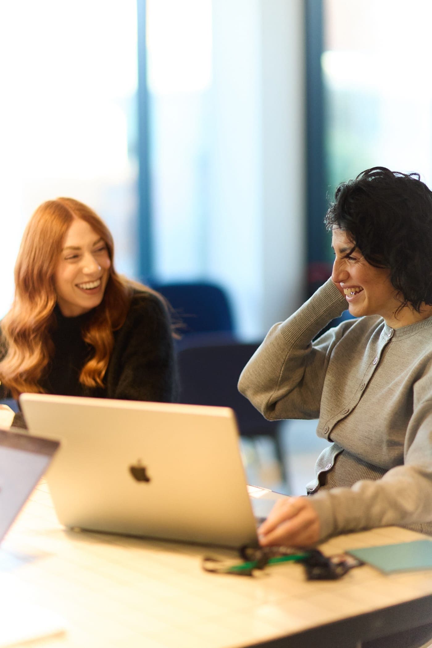 Two women smiling and talking while working on laptops in a bright office with large windows.