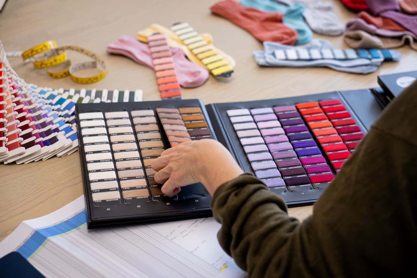 Person browsing colour thread swatches on a table covered with yarn samples, striped socks, and measuring tape.