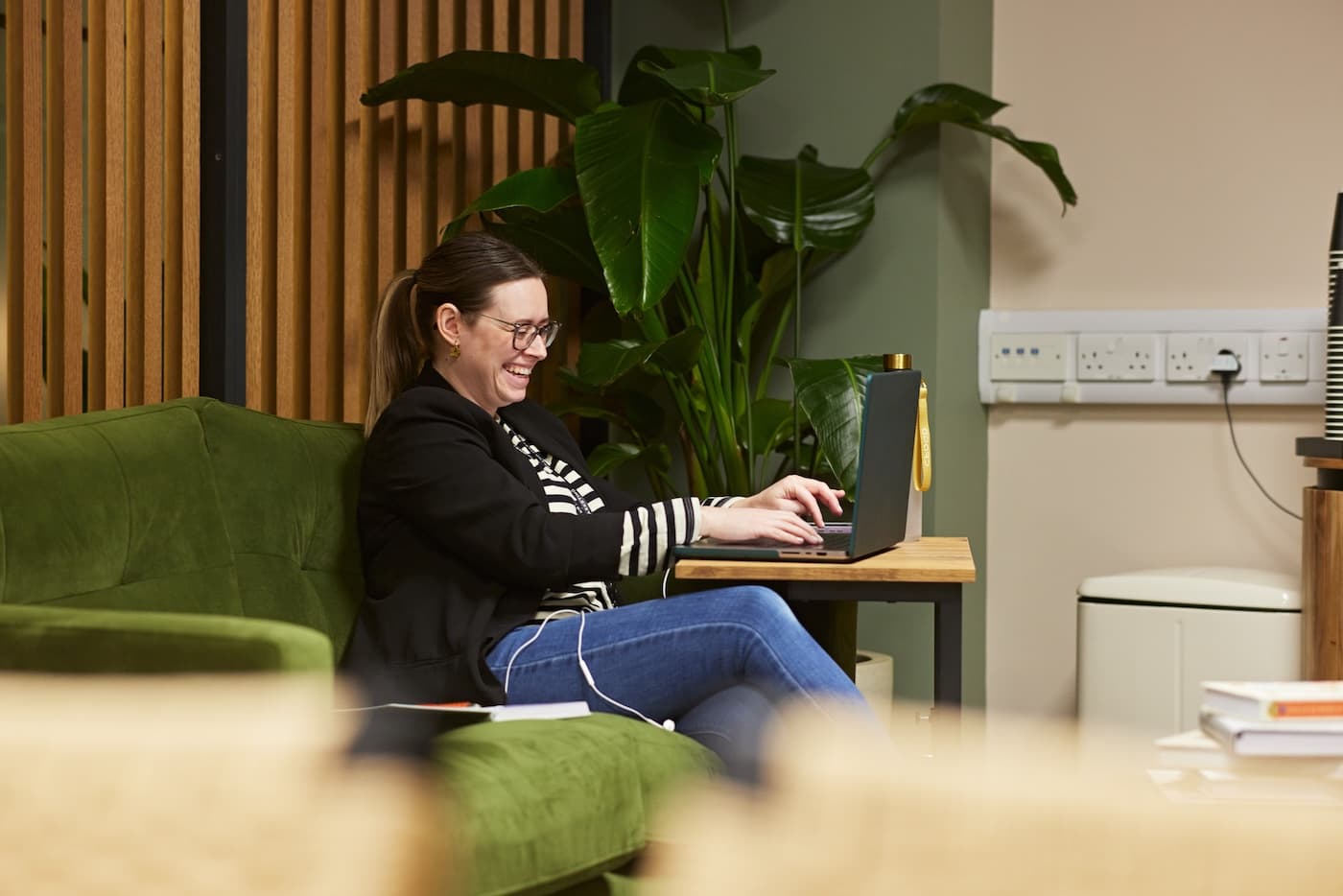 Woman sitting on a green sofa in a relaxed office setting, smiling while working on a laptop, with a large plant and power outlets in the background.