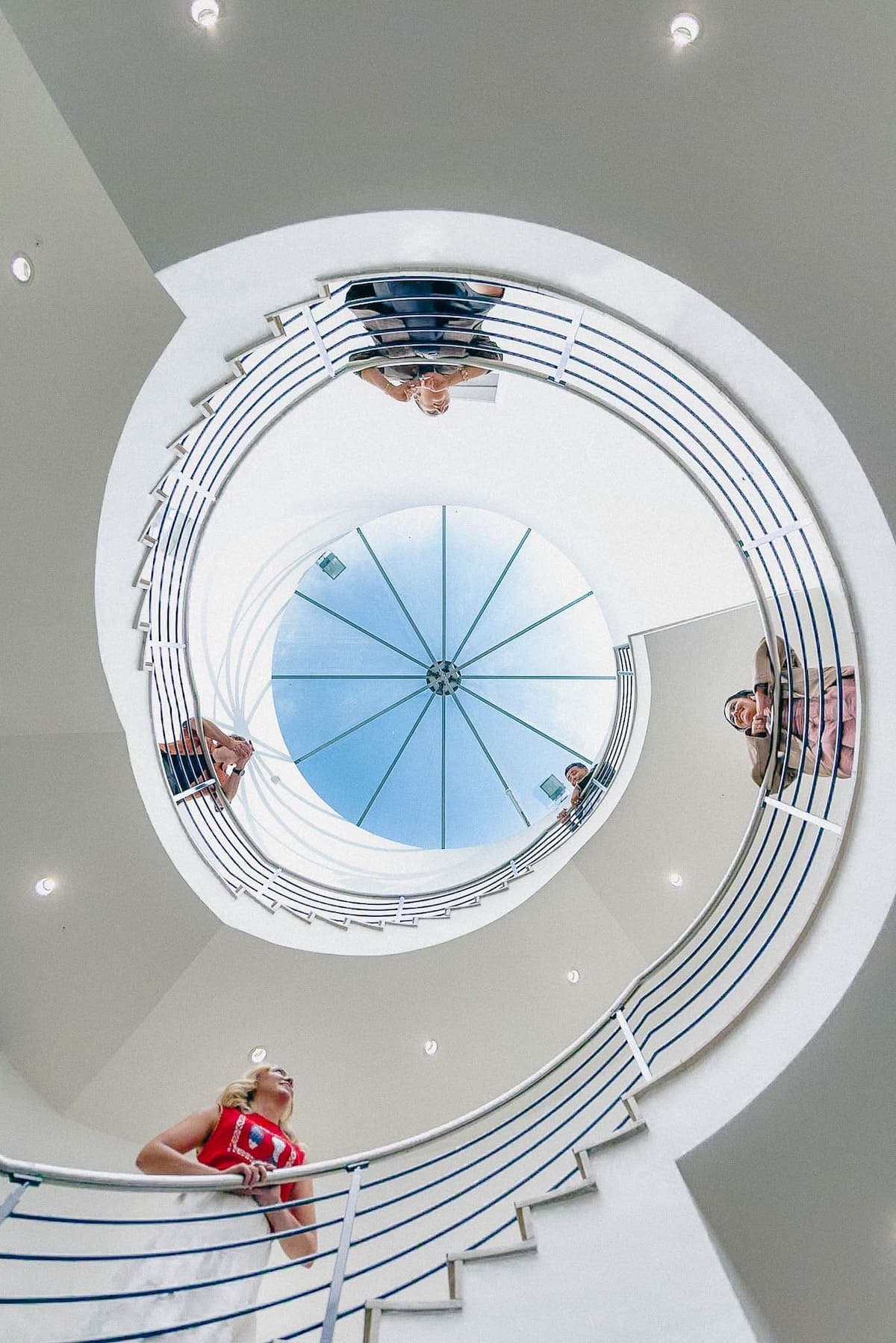 Upward view of people leaning over a spiral staircase railing beneath a glass dome ceiling.