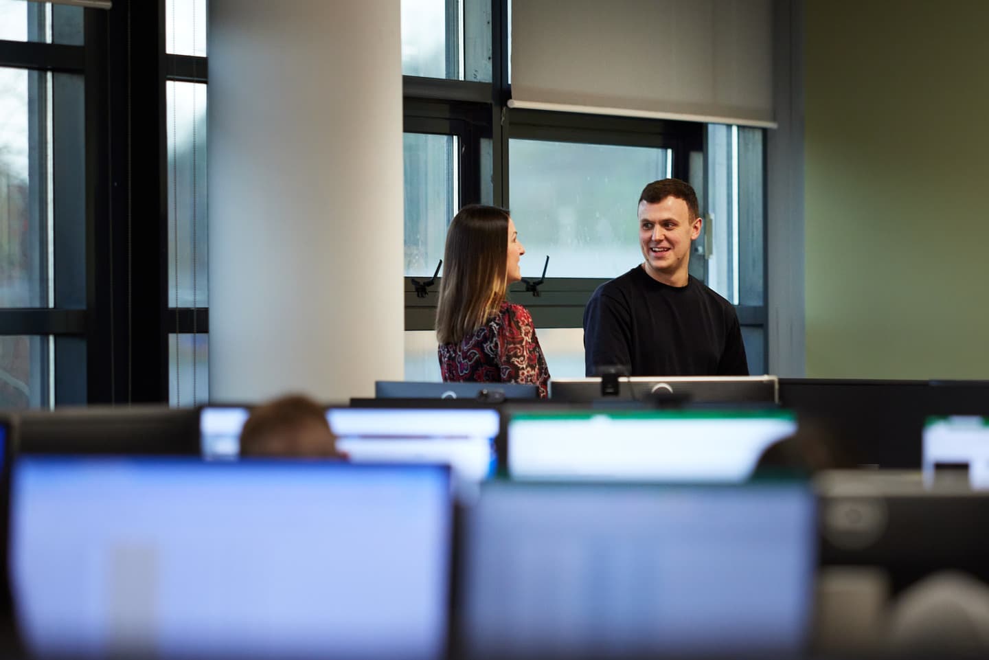 A female and male colleague are stood together by the windows in the office, smiling and engaged in conversation. Computers with blurred screens are visible in the foreground.